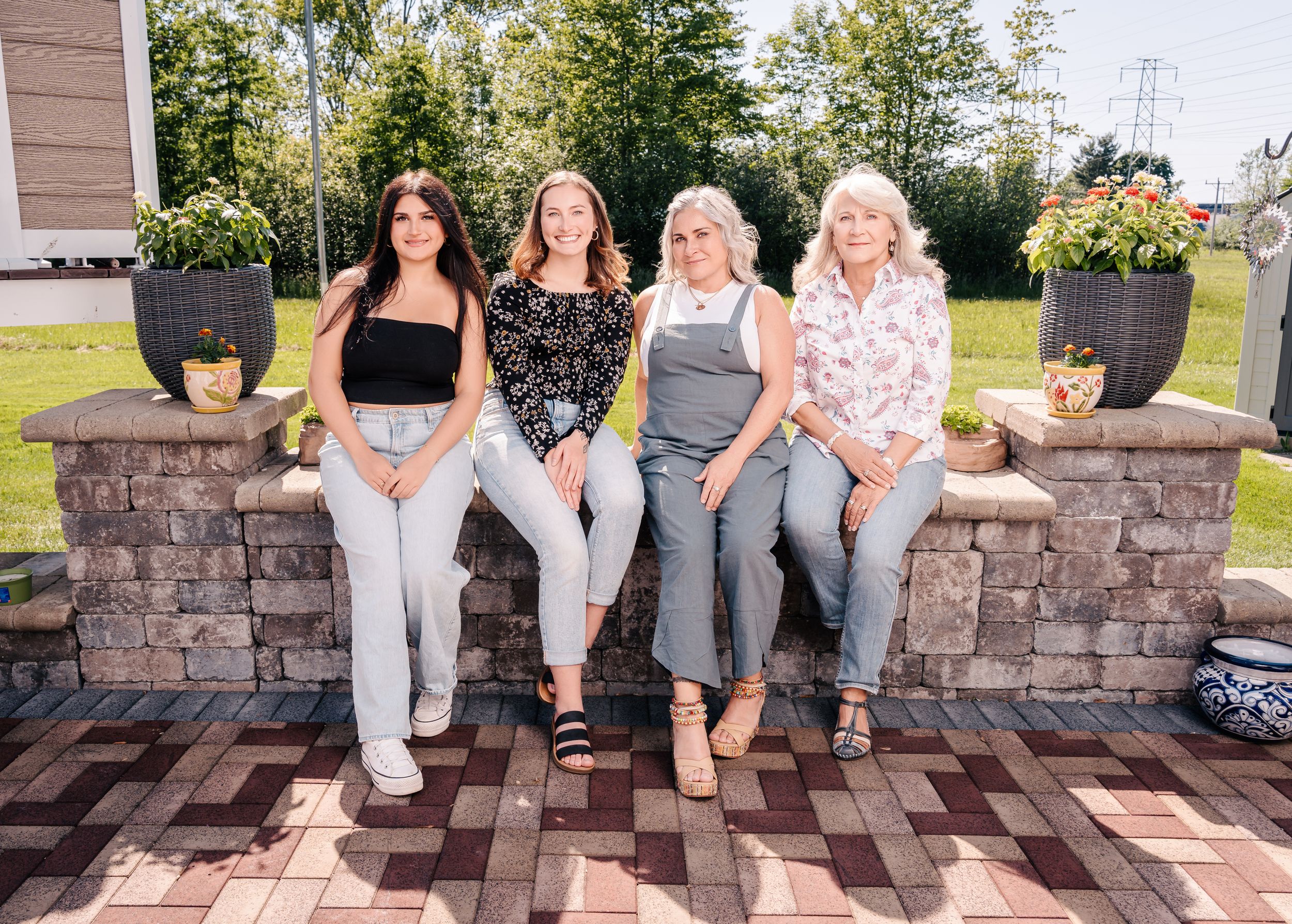 Four women sit casually together on a stone wall with planters on a wooden deck.