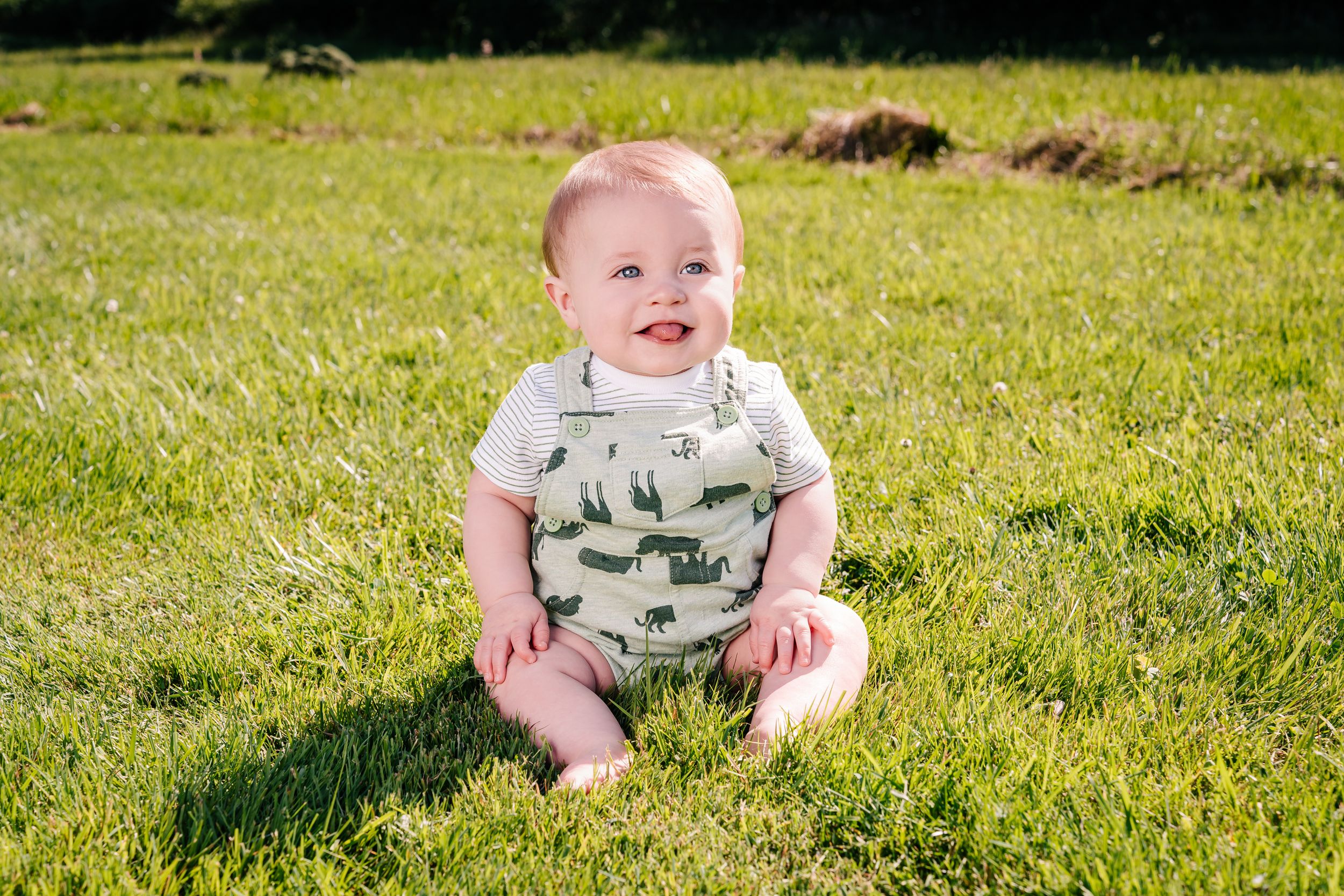 Happy baby wearing dinosaur print overalls sits on sunny green grass in a garden.