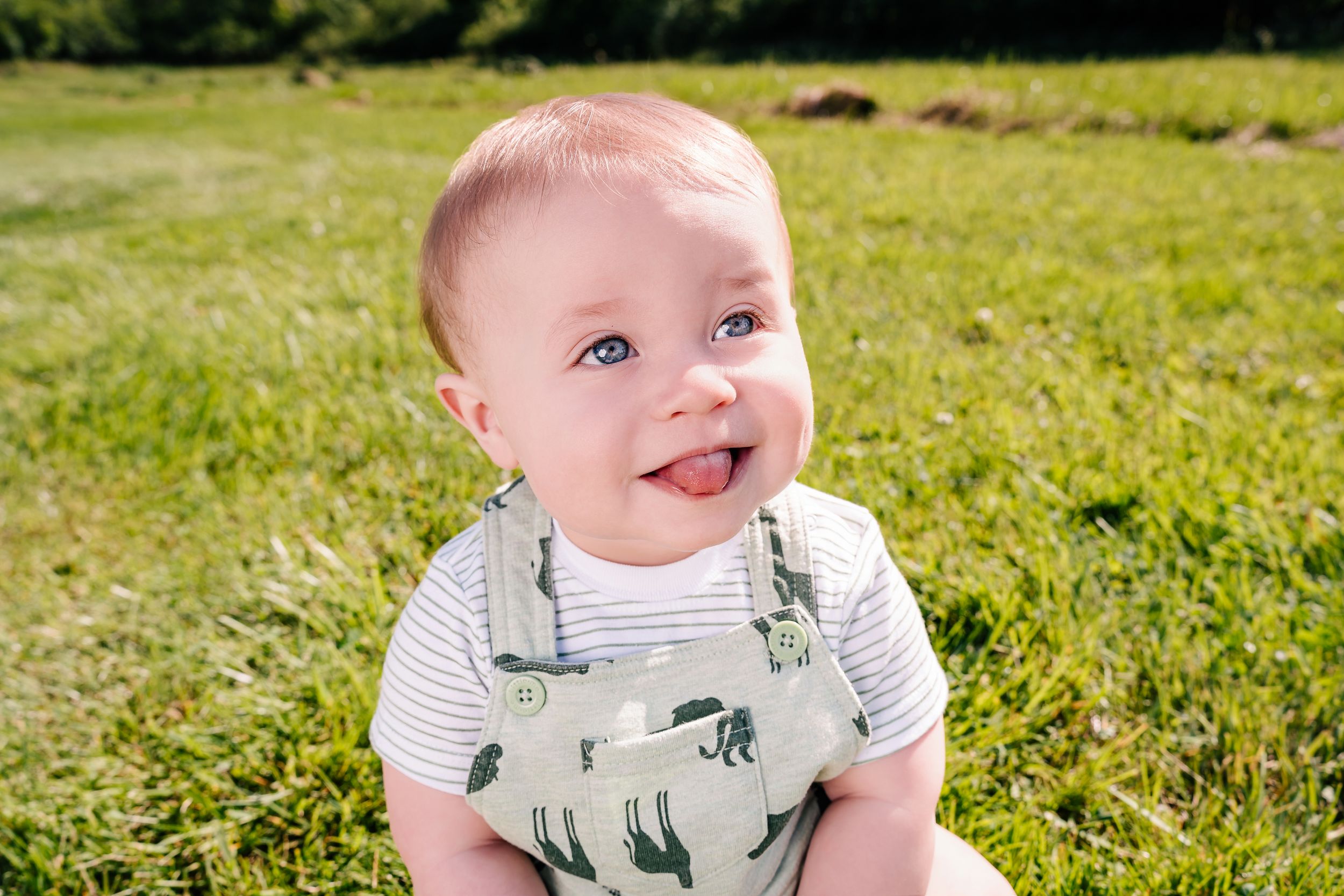 Joyful baby in dinosaur print outfit laughs while sitting outdoors on lush grass.