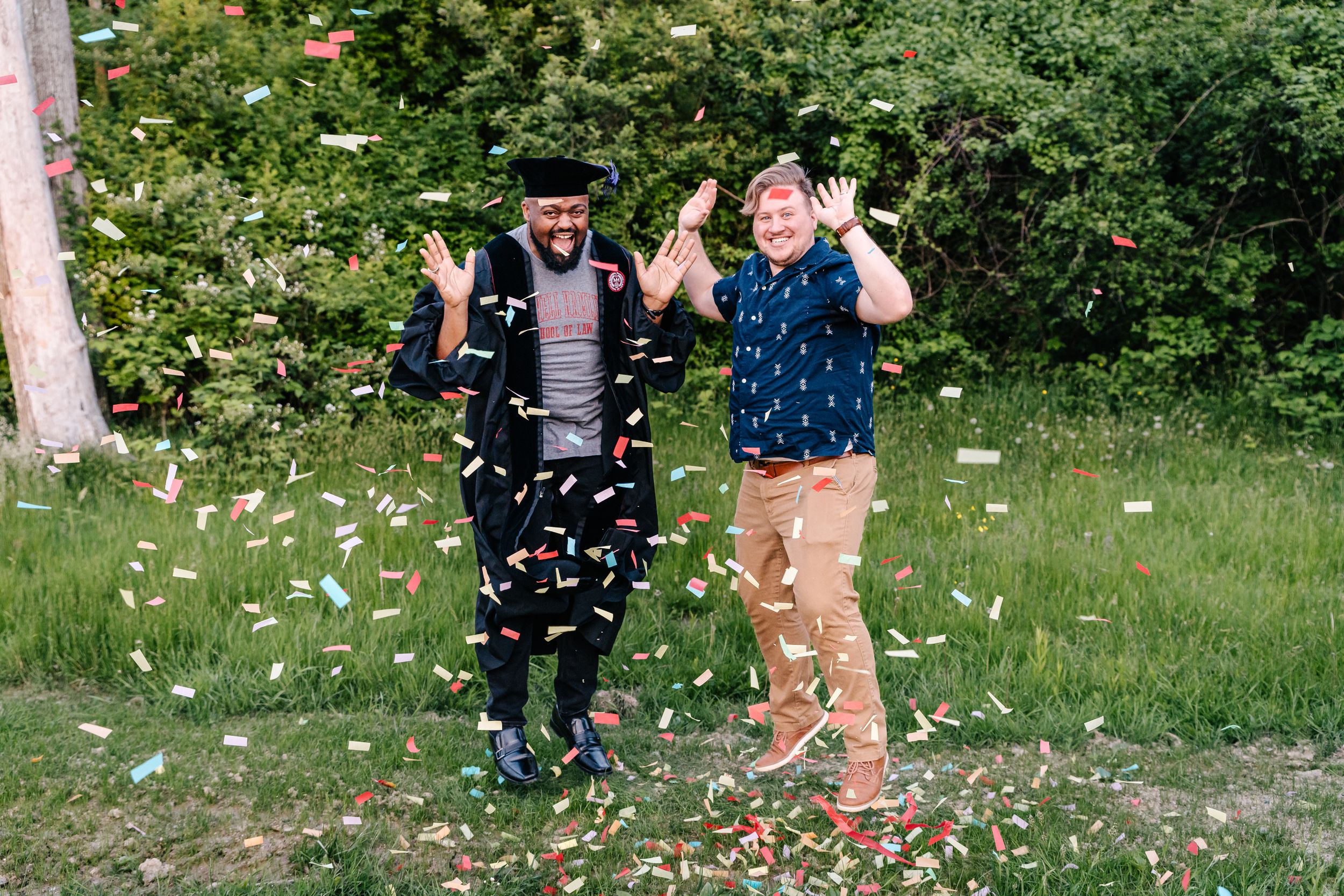 Two people celebrate joyfully as white confetti falls around them in a green garden setting.