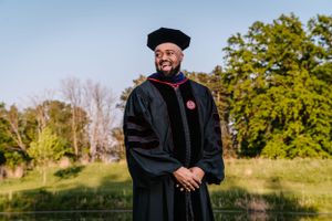 Graduate in traditional black academic regalia and cap standing outdoors near a pond.
