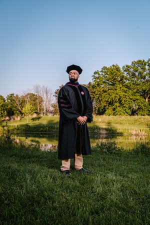 Academic in full graduation regalia standing on grass with a pond and trees in the background.