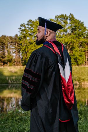 Side view of graduate in doctoral regalia with red accents against a natural outdoor backdrop.