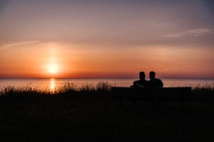 Two silhouettes sit together watching a vibrant orange sunset over calm waters.