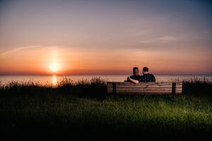 A couple sits on a bench overlooking the ocean during a stunning orange sunset.