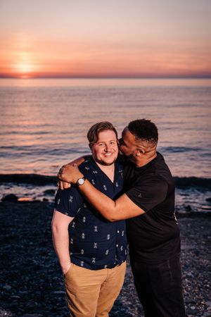 Two people embracing on a beach at sunset with orange sky and ocean waves.