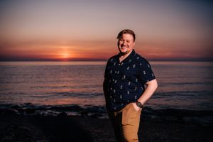 Person in navy patterned shirt smiling on rocky beach during golden hour sunset.