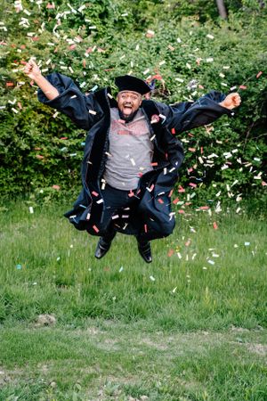 Person in black jacket jumps with joy as confetti falls in a garden setting.