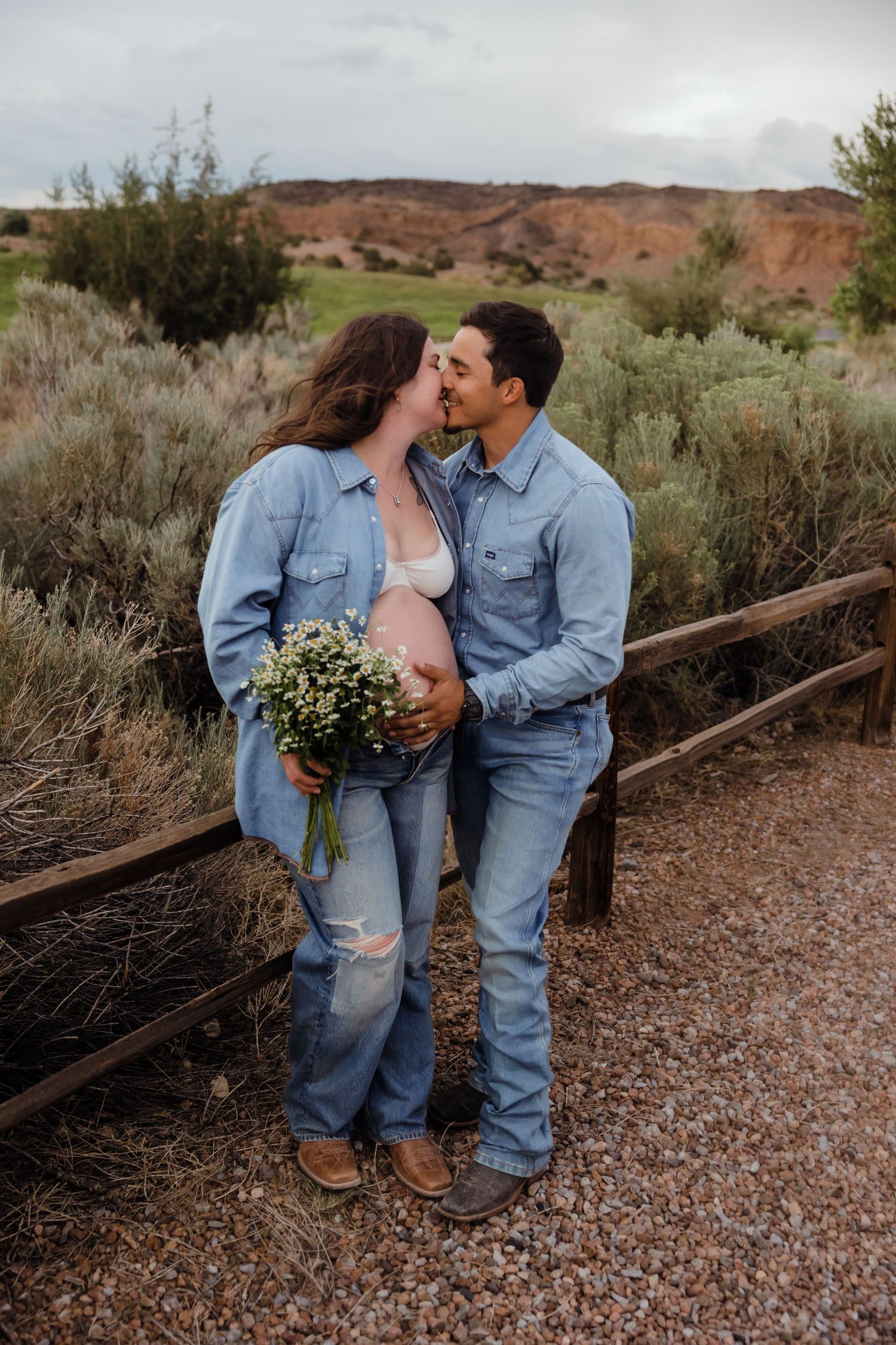Pregnant couple in matching denim kissing outdoors, woman holding wildflower bouquet at fence.