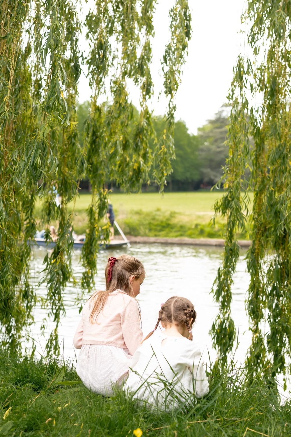 Two girls sit by a peaceful river Cam under weeping willow branches in a lush green park in Cambridge, UK