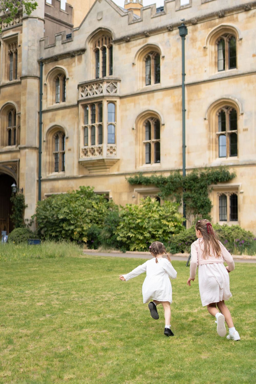 Two girls in white dresses running across a green lawn in front of Trinity College, Cambridge, UK