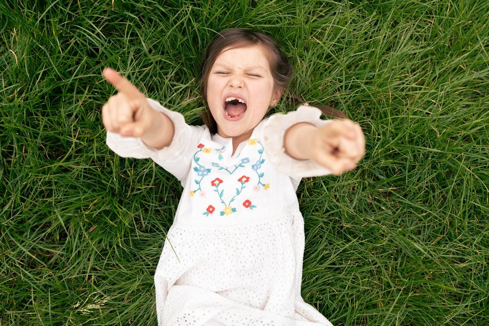 Happy young girl lying on green grass with arms outstretched, smiling and pointing upward in a white dress. Jesus Green, Cambridge, UK