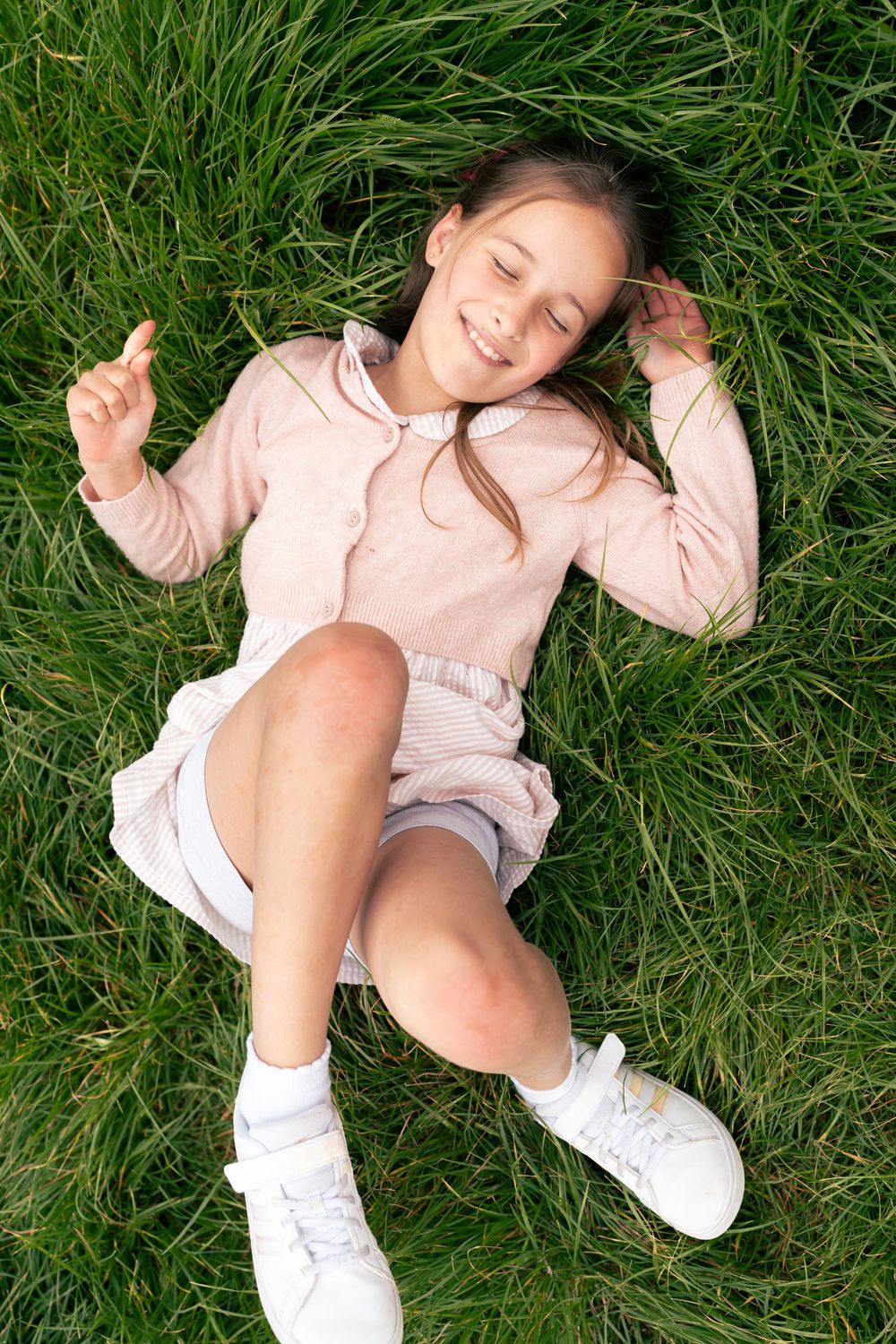 Young girl relaxing on green grass wearing a pink dress, purple shorts, and white sneakers in an outdoor setting. Jesus Green, Cambridge, UK
