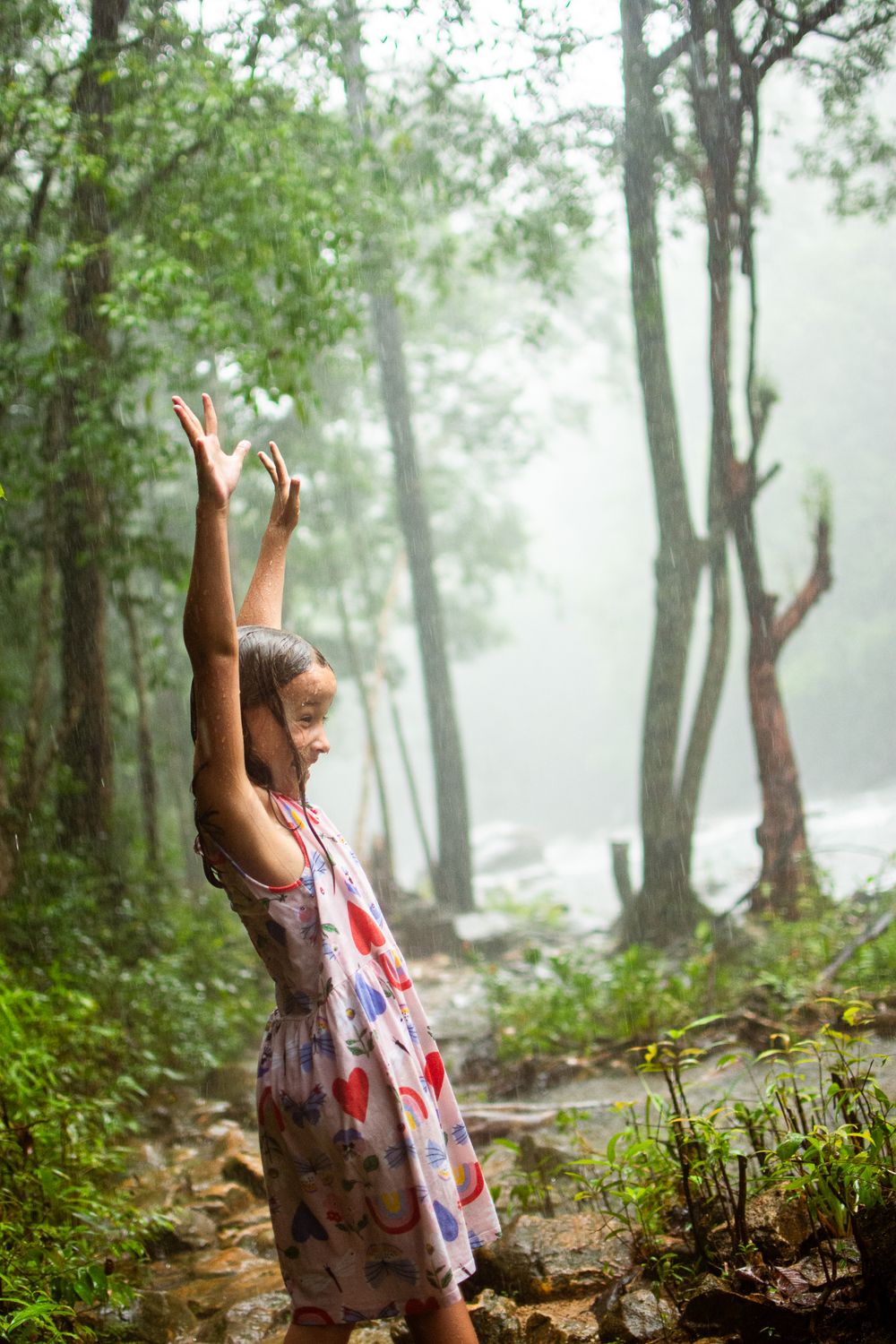 A little girl in a floral dress stands with arms raised joyfully on a misty forest trail surrounded by green trees in Thailand, Koh Chang
