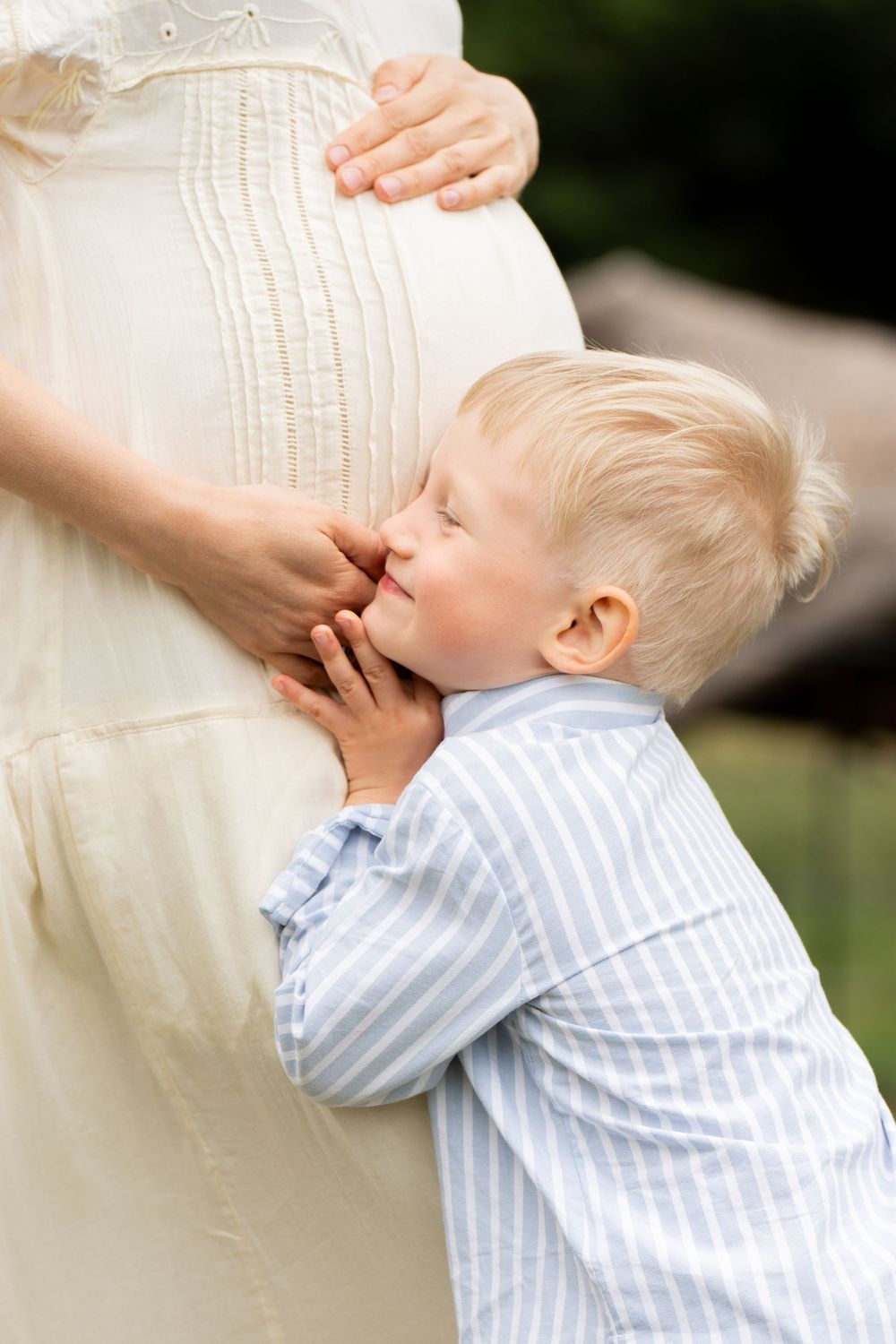 A young child in striped shirt hugs and kisses a pregnant belly while wearing a sweet expression outdoors. London UK