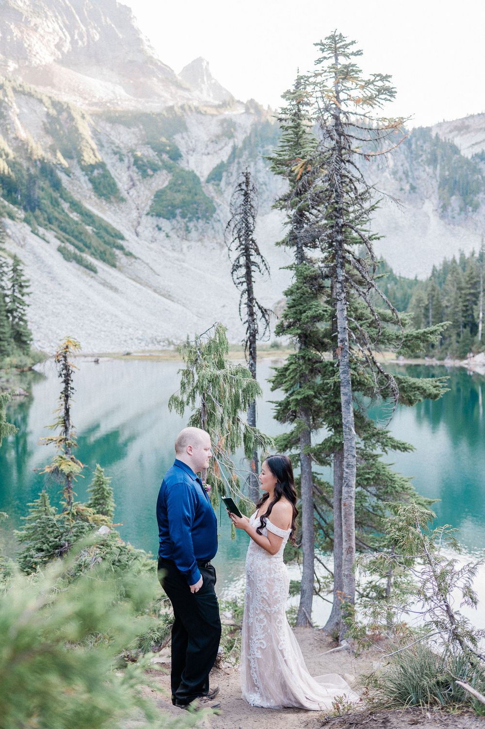 Bride and groom exchange vows within Mt. Rainier Park with evergreen trees and snow-capped peaks reflecting in turquoise alpine waters.