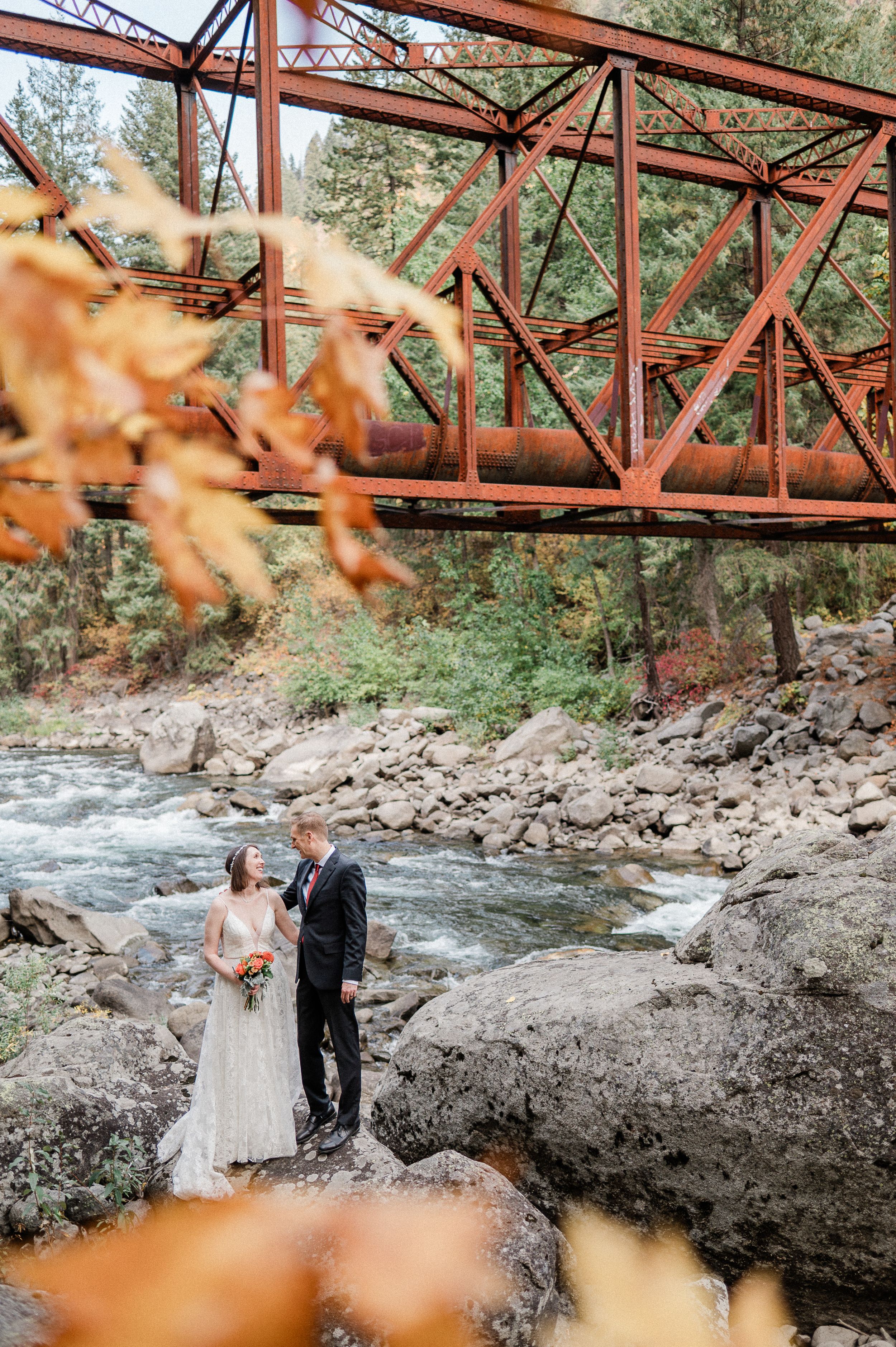 Bride and groom featuring a red metal bridge with autumn leaves and rocky riverbed in the background during their Washington elopement.
