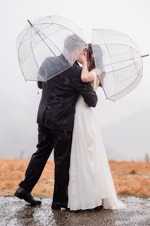 bride and groom kissing while raining and holding clear umbrellas