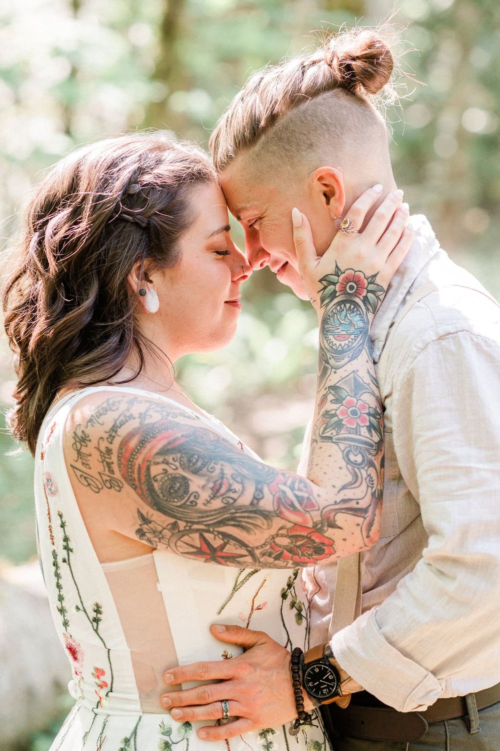 two brides forehead to forehead, one with tattoo arms, embrace in the woods.