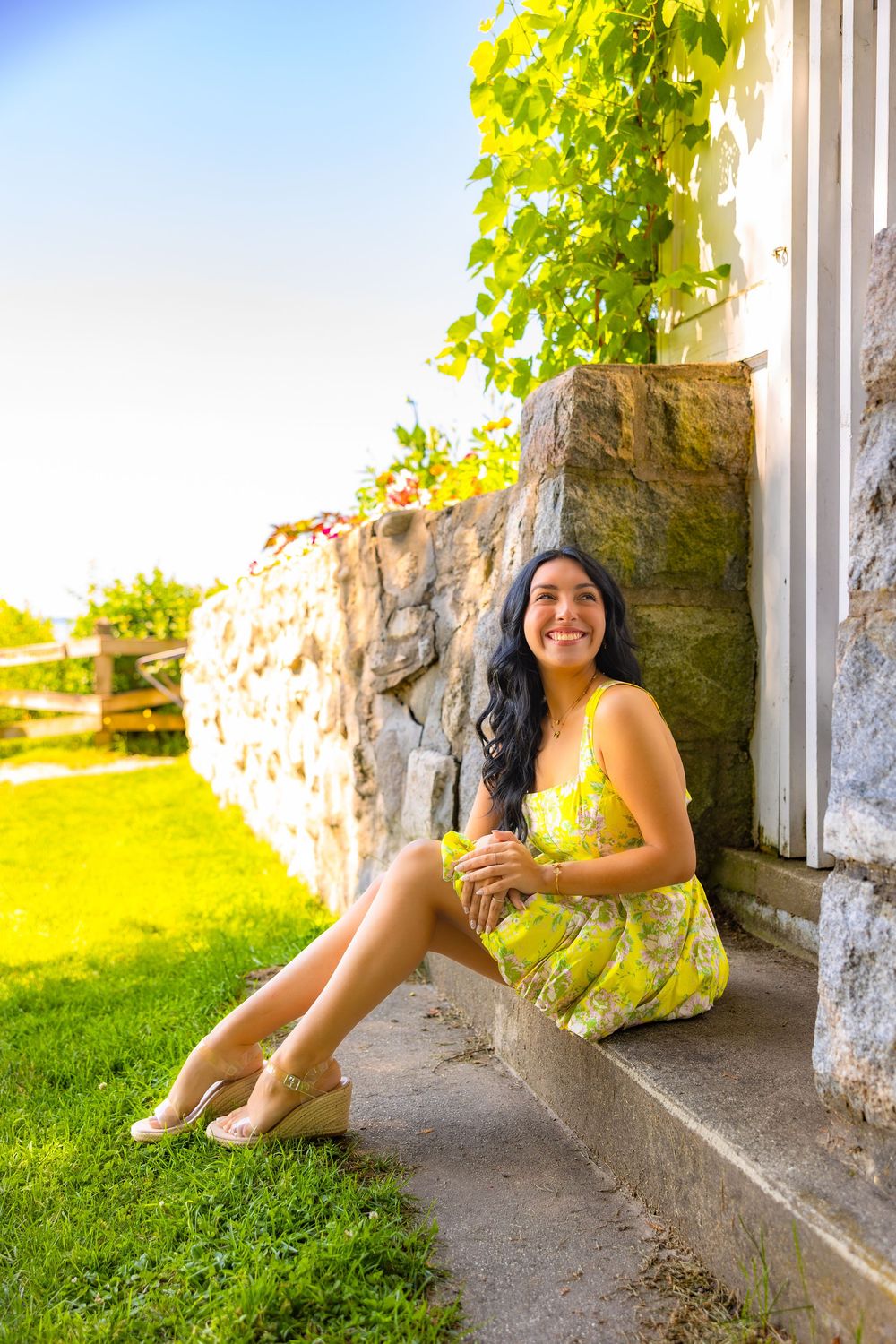 A person in a yellow floral dress sits on stone steps on a sunny day with green grass and vines in the background.