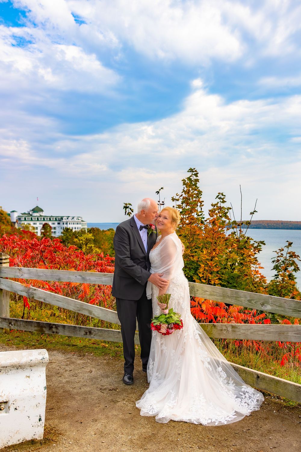 Couple stands on scenic overlook with vibrant red autumn foliage and waterfront view on a bright cloudy day.