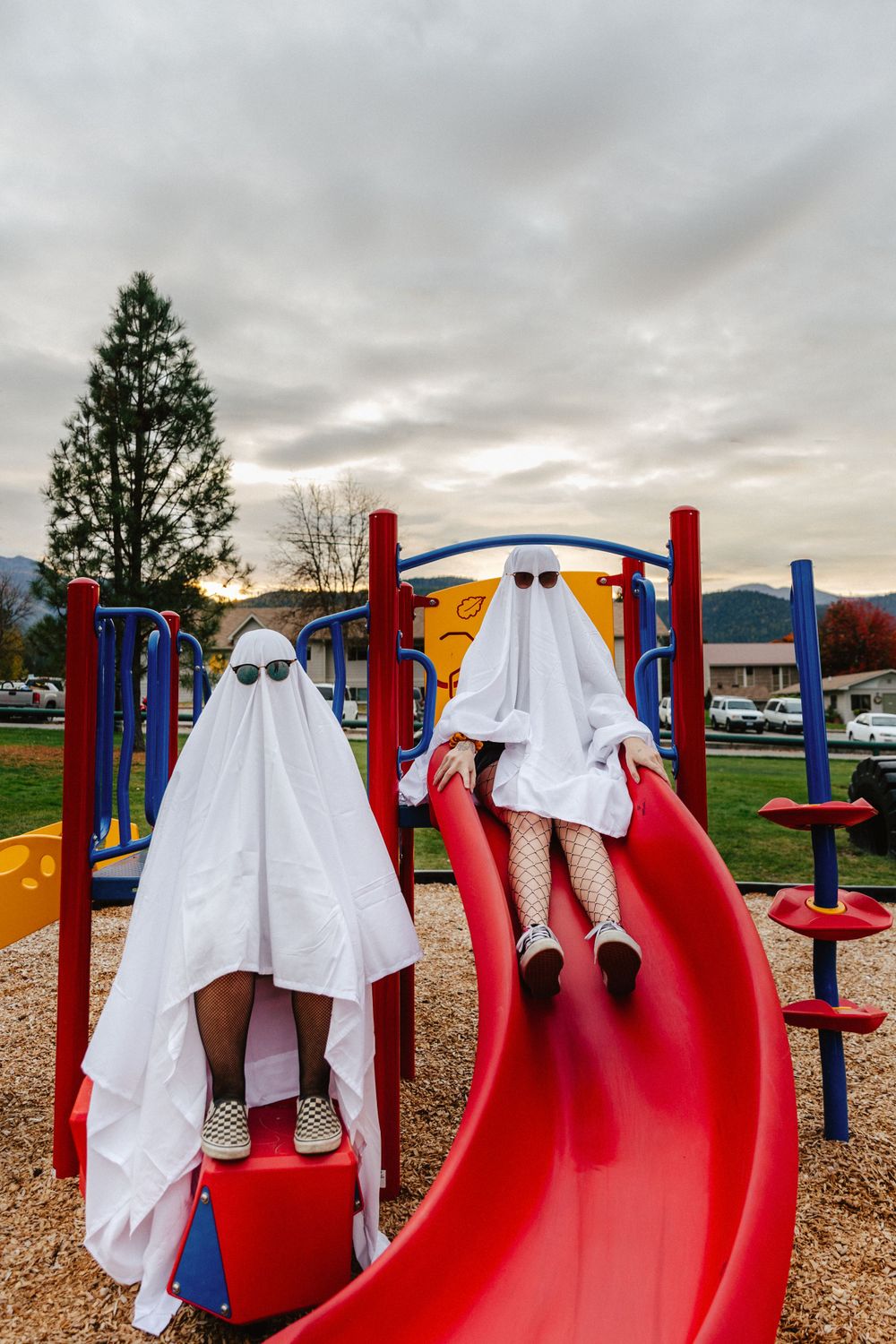 Two ghostlike figures wearing white sheets and sunglasses play on a red playground slide on a cloudy day.
