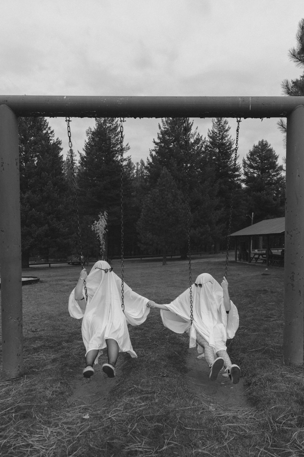 Two figures in ghost costumes made from white sheets swing together on playground equipment in a black and white photo.