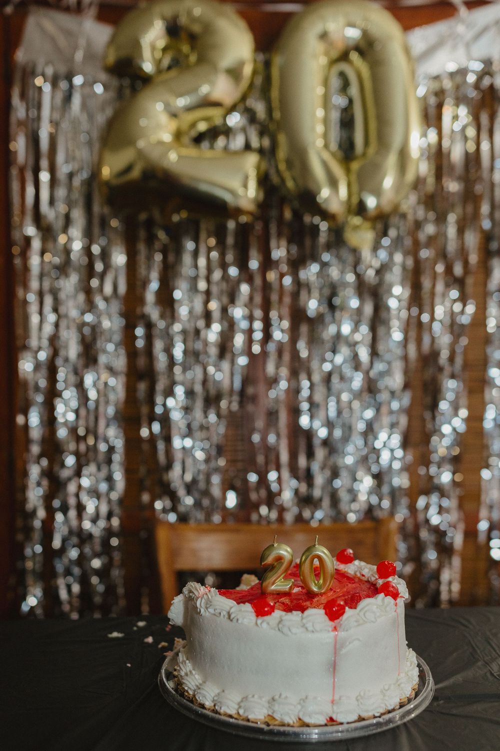 White birthday cake with red accents and number 20 topper sits against silver tinsel backdrop with gold balloon numbers.