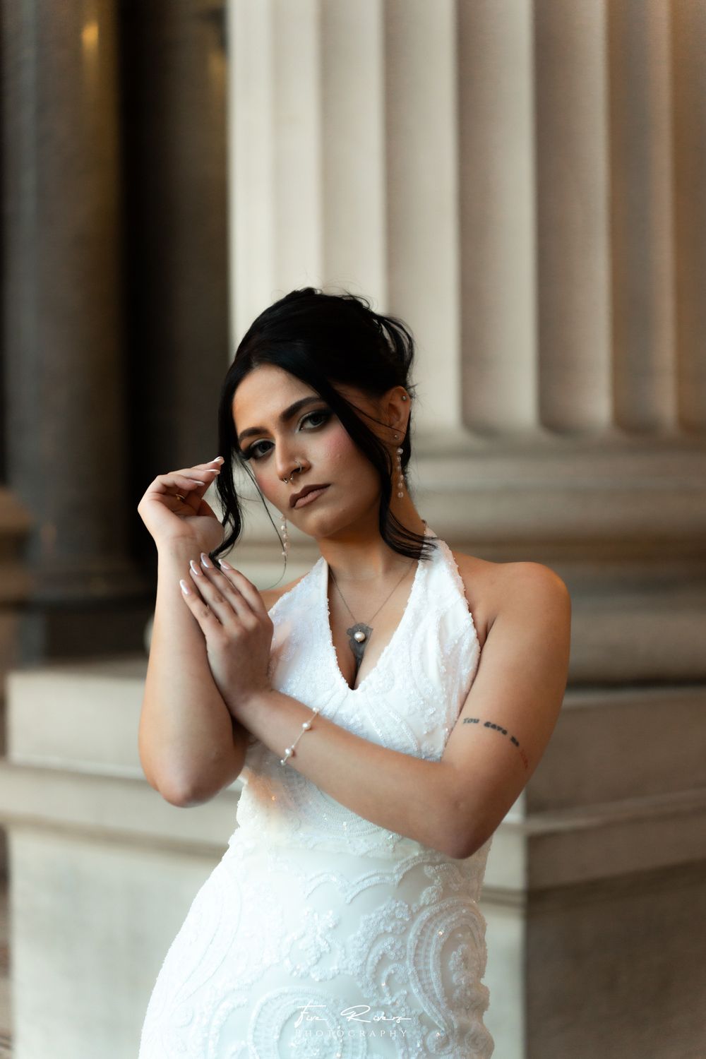 A bride in an elegant white lace gown poses against classical stone columns, adjusting her delicate jewelry.
