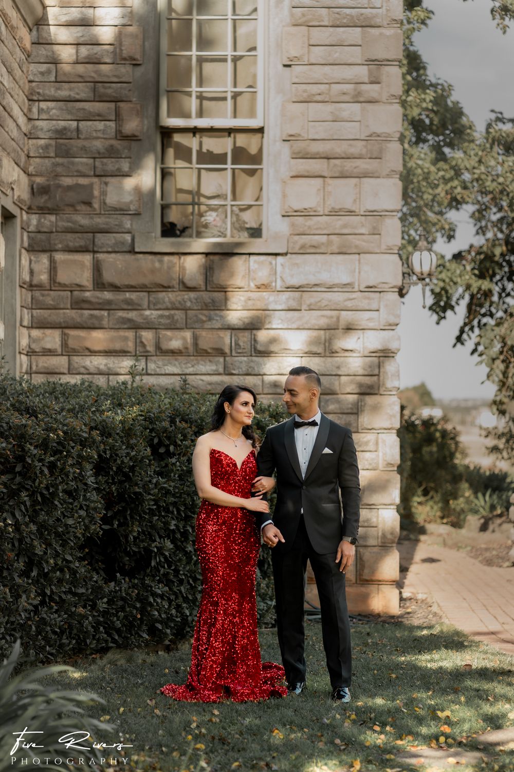 Elegant couple poses by stone building, with sparkly red gown and black tuxedo creating a romantic formal portrait.