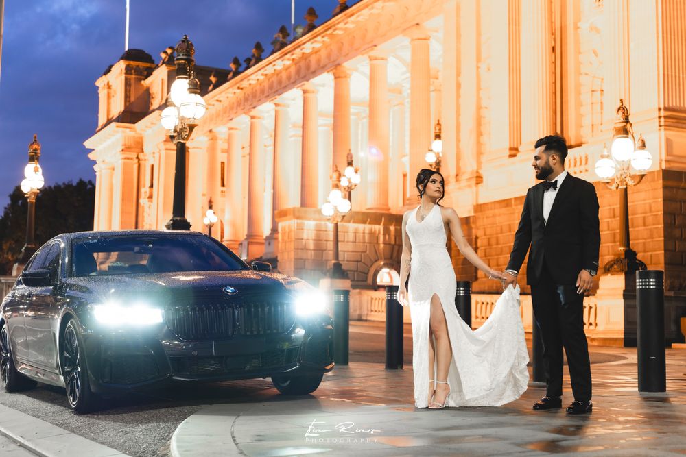 A couple stands by a luxury BMW car at night in front of an illuminated ornate historical building.