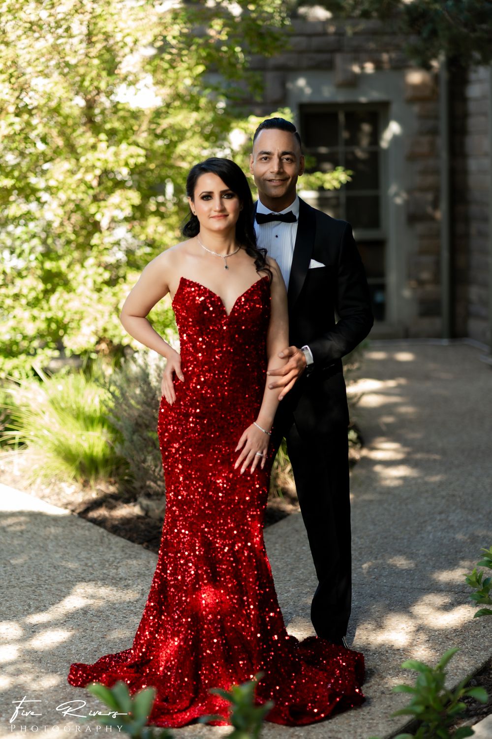 A couple poses outdoors in elegant attire, with a stunning red sequined gown and classic black tuxedo.
