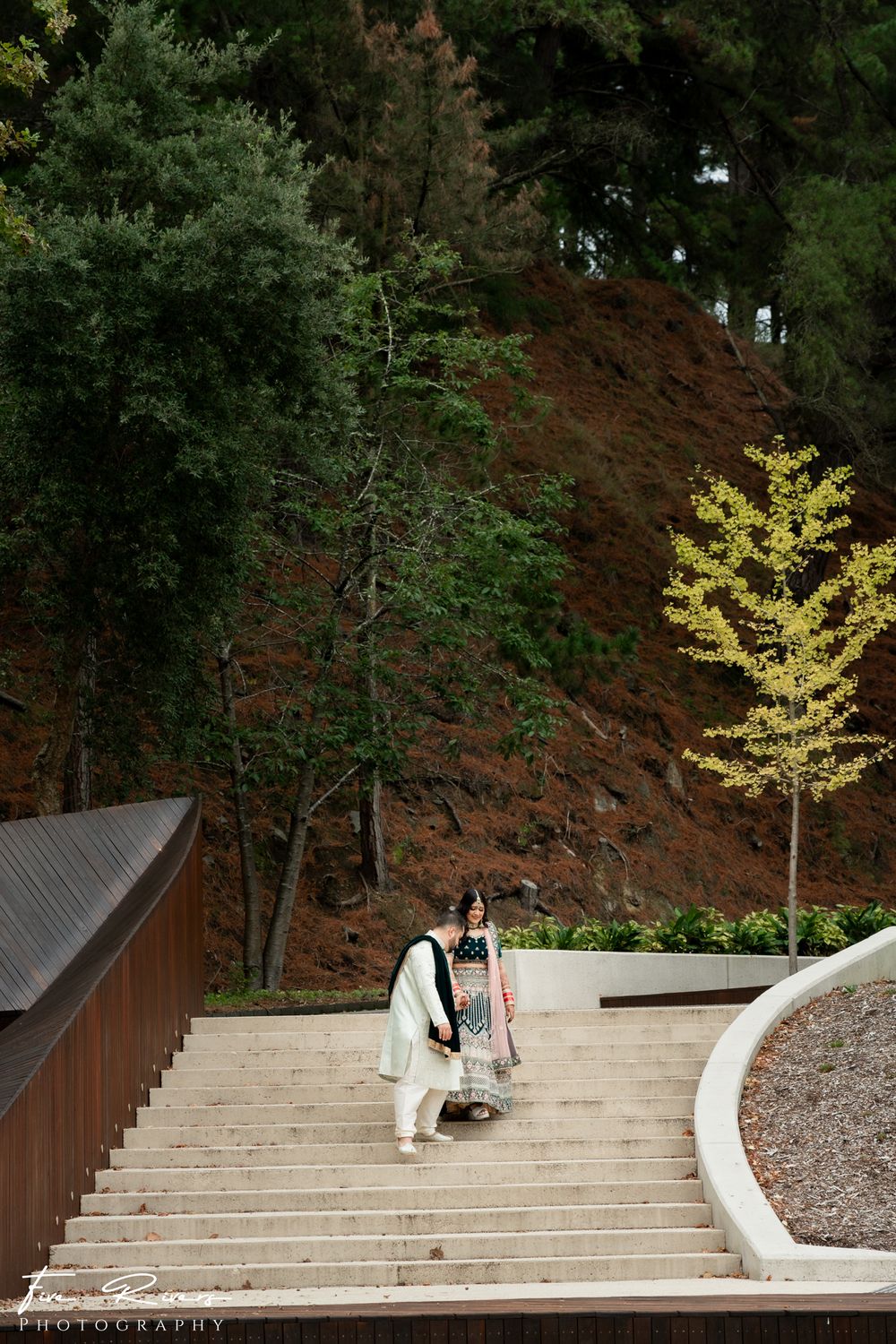 A person walks up concrete steps alongside a curved wall and railing surrounded by lush trees and red cliffs.