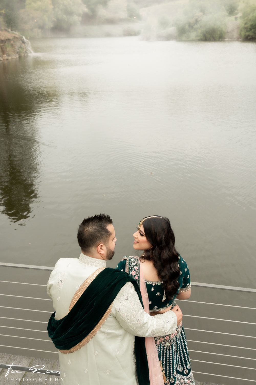 A couple embraces while sitting on metal railings by a misty lake during an outdoor photoshoot.