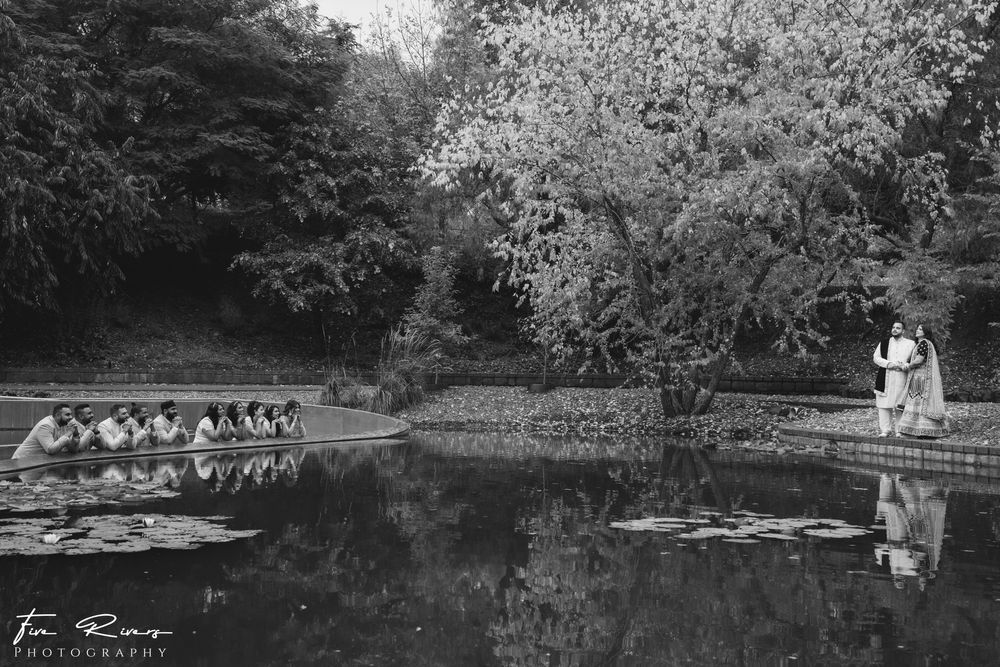 Black and white reflection of wedding couple standing by tranquil pond with rowboats lined up on the shore.