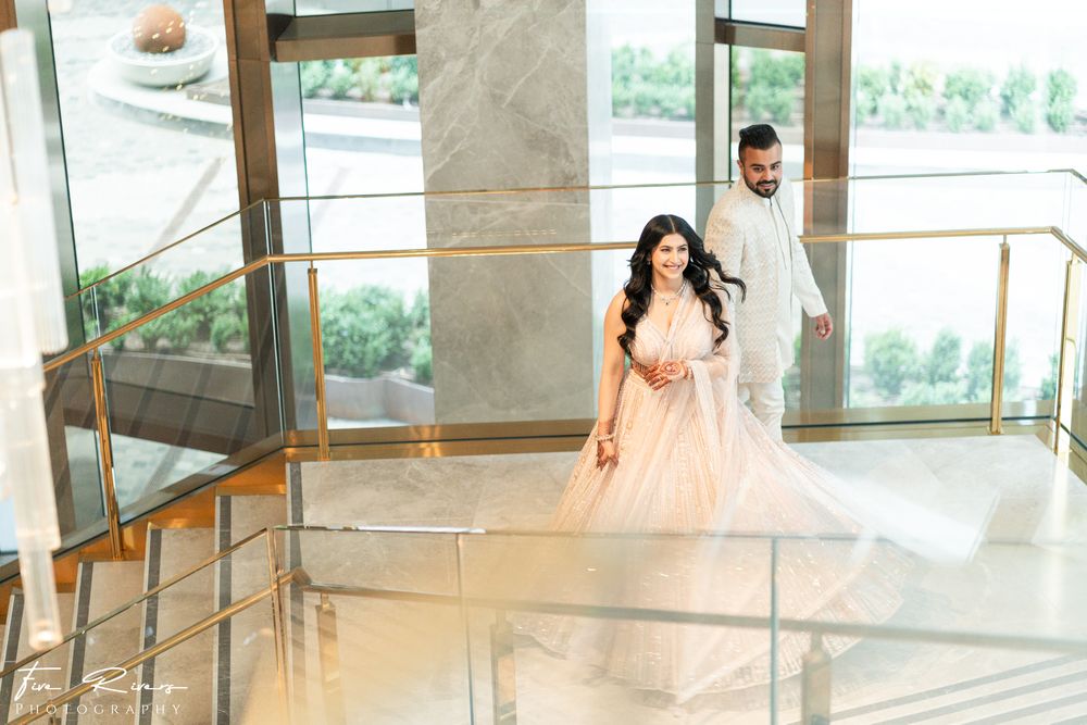 A couple in formalwear descend an elegant marble staircase in a modern building with glass walls and natural light.