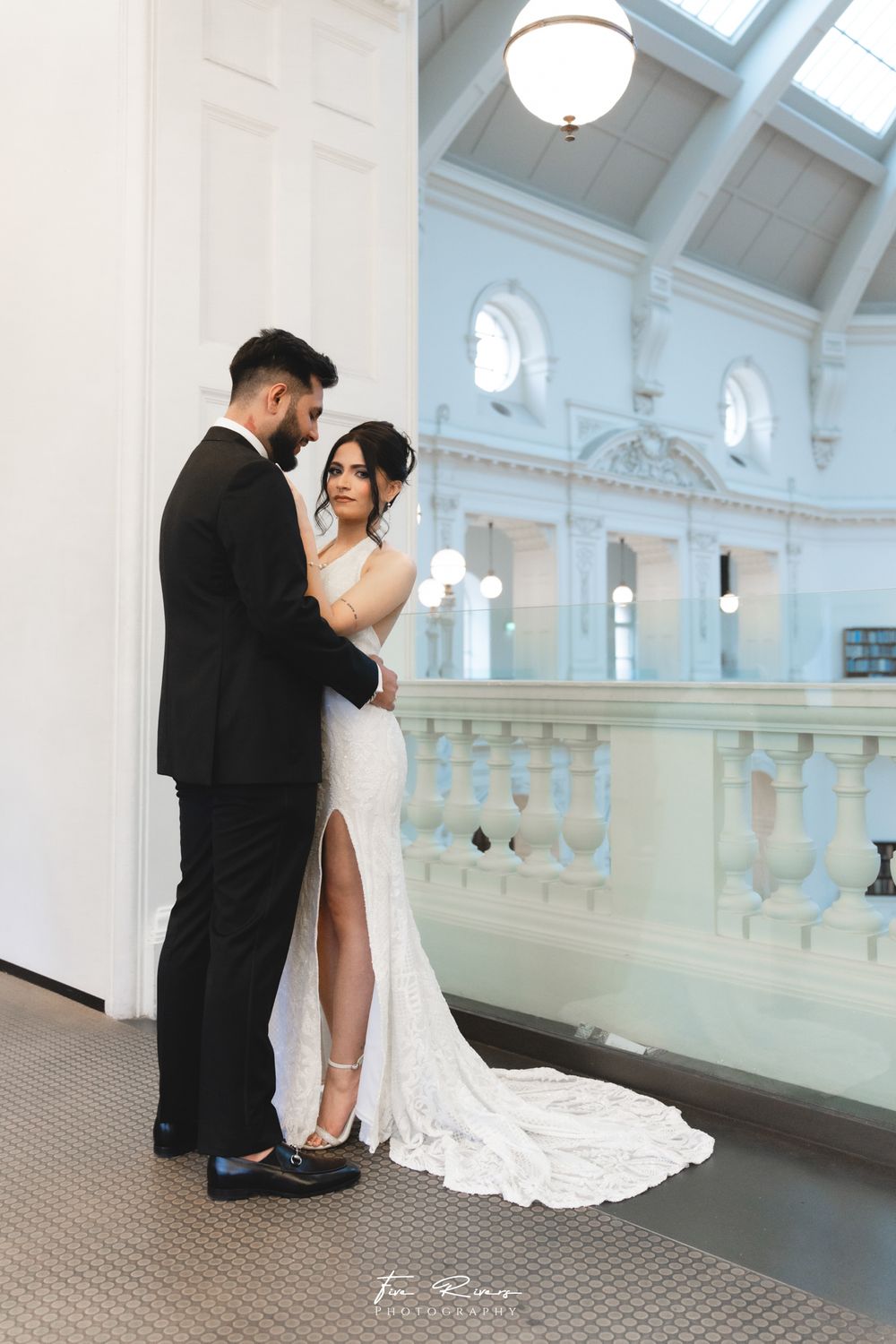 Couple in wedding attire embrace in an elegant venue with high ceilings, arched windows and white architectural details.