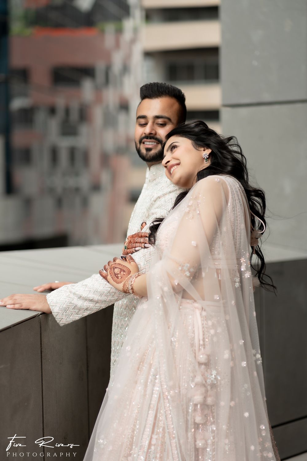 A couple in traditional South Asian wedding attire embrace and smile while standing on a balcony in an urban setting.