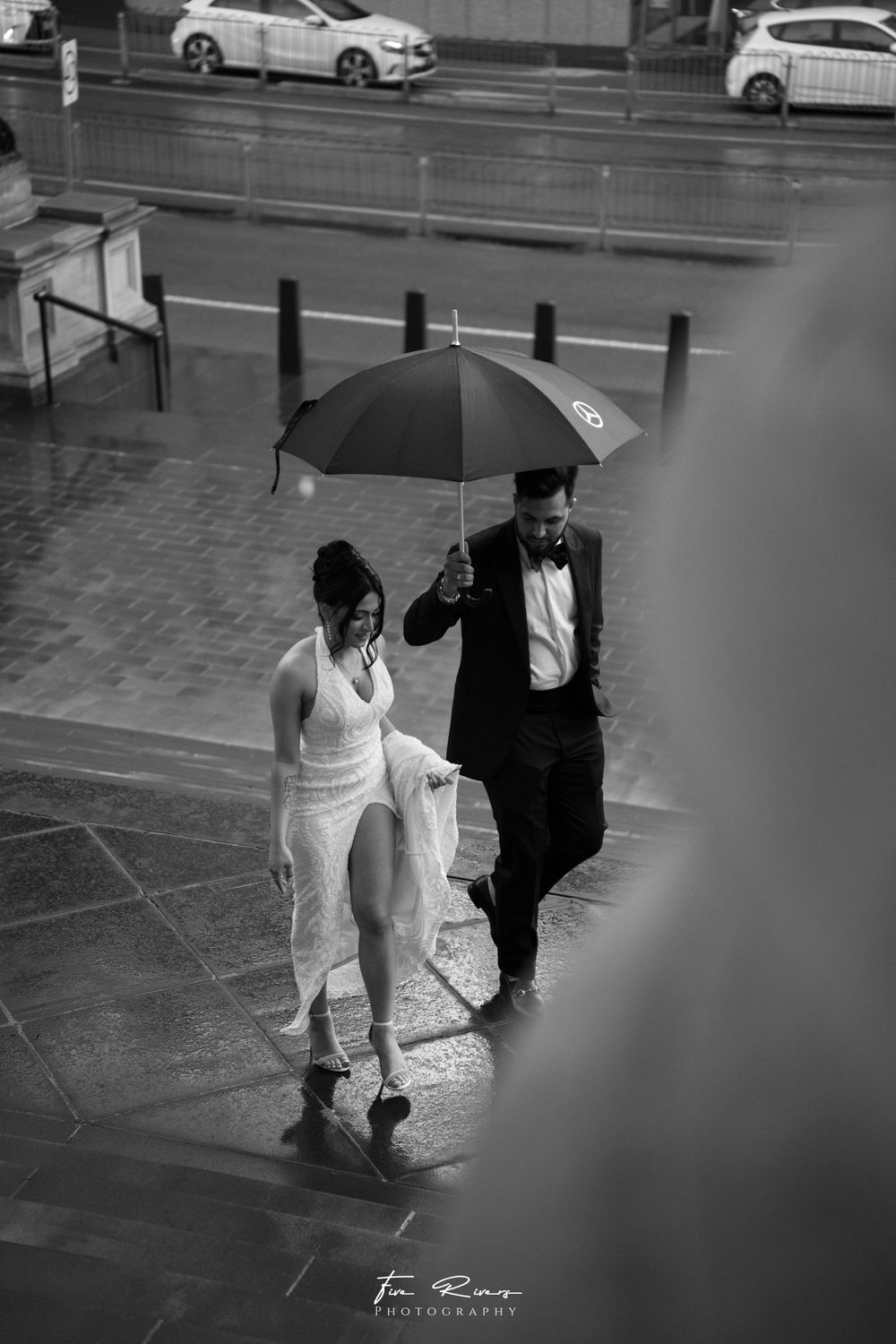 A couple in wedding attire walks under a black umbrella on a rainy day in a black and white street scene.