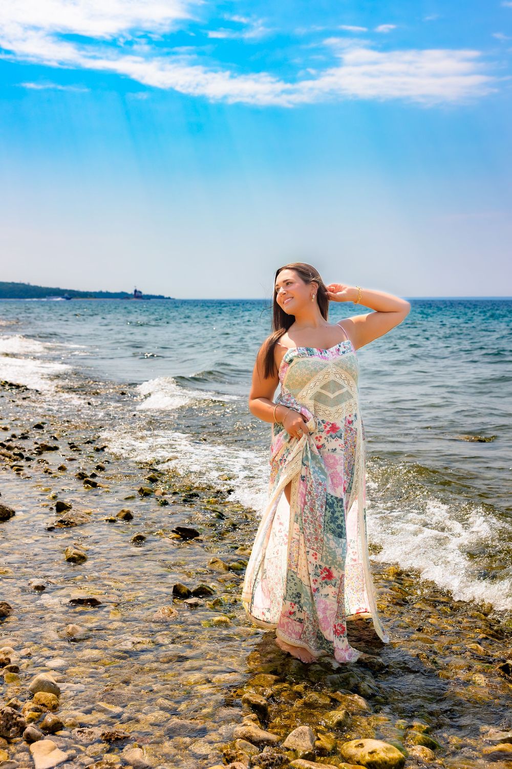 A person in a floral dress poses dramatically by the rocky shoreline on a bright sunny day at the beach.