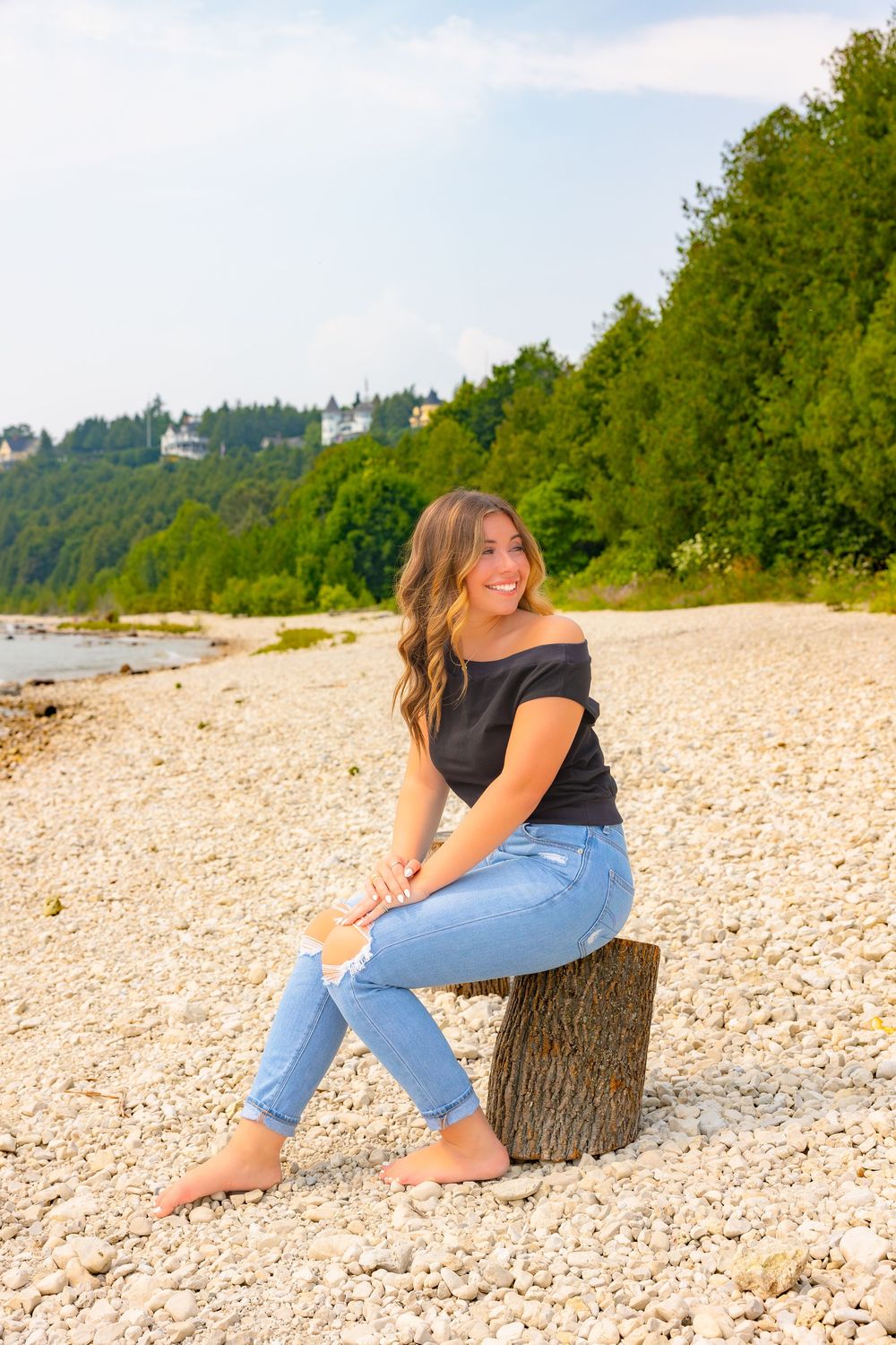 Person in black top and jeans sits barefoot on driftwood log at rocky beach with forested hillside in background.