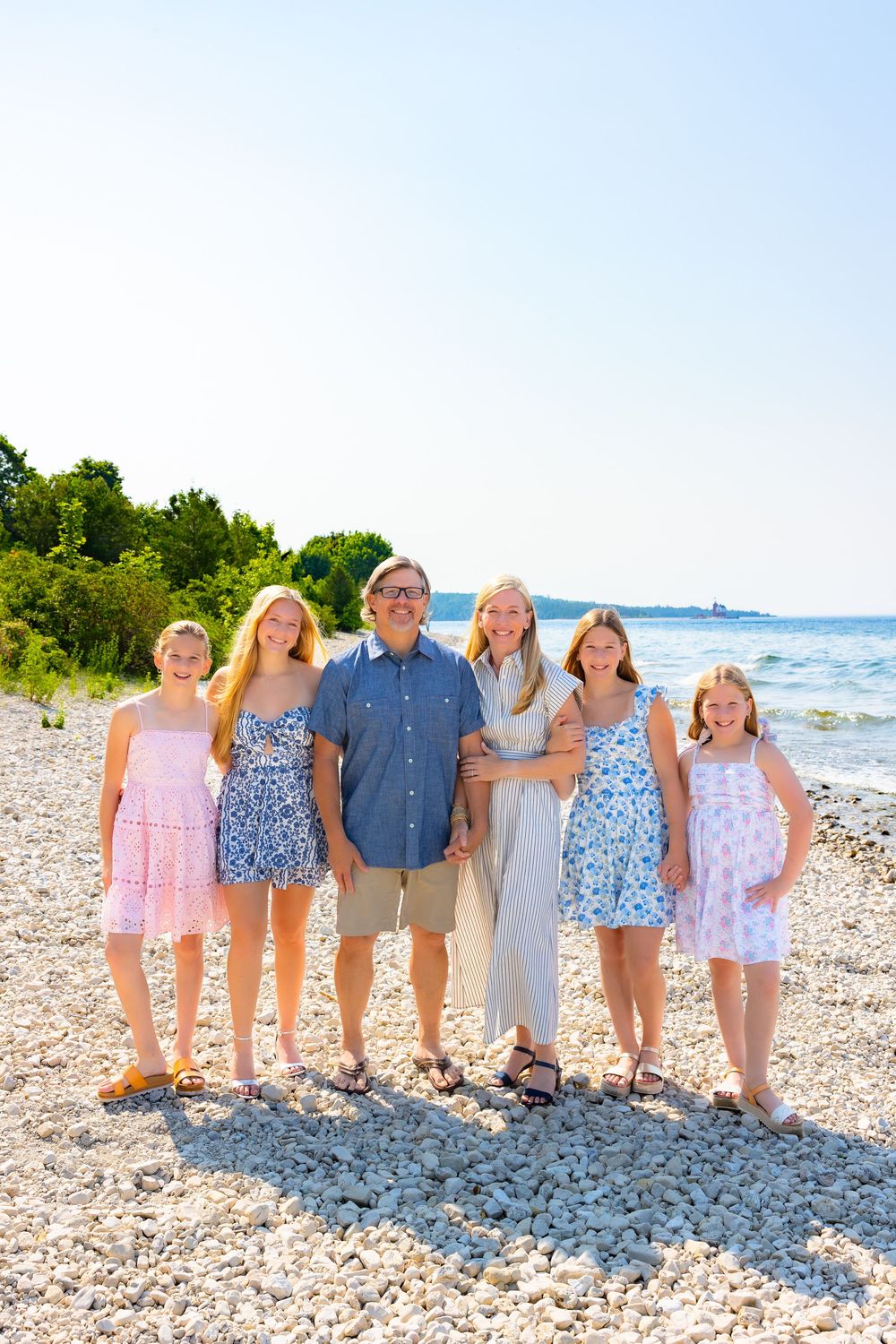 A group stands together on a rocky beach shoreline with blue water and green trees visible in the background.