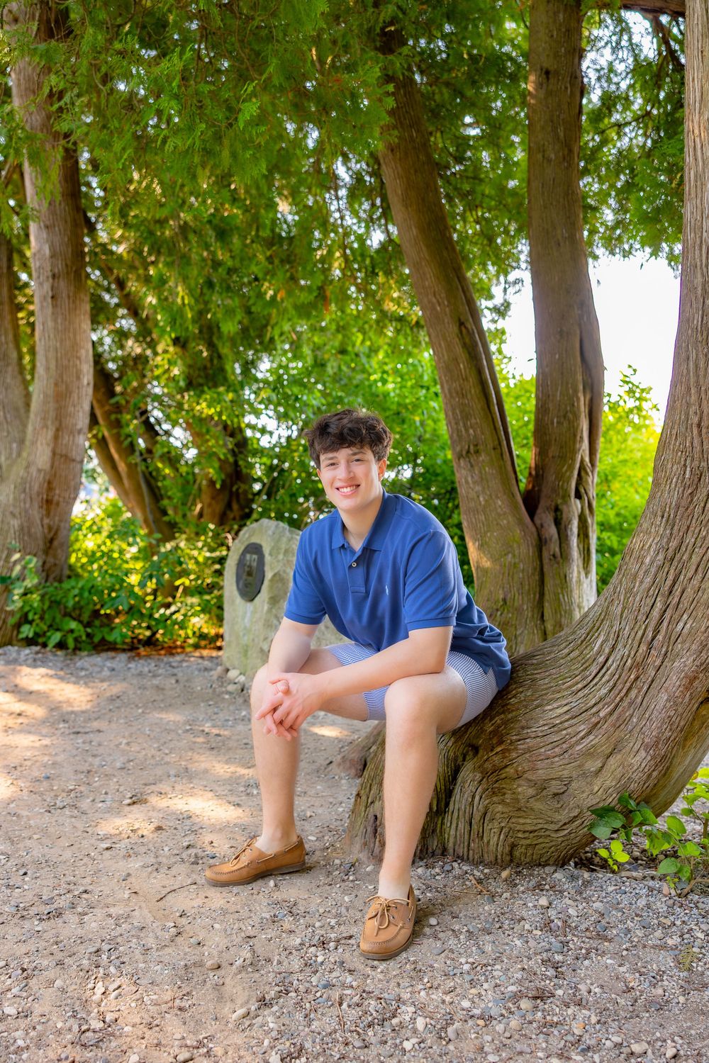 Someone sits on a gravel path beneath curved tree trunks wearing blue shorts and brown boots.