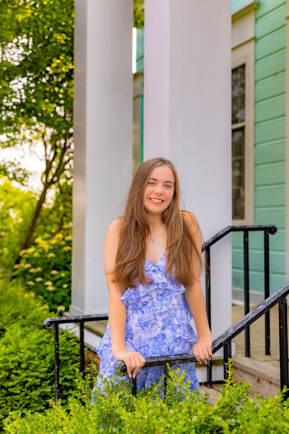 A person in a blue floral summer dress stands on the steps of a white-columned building surrounded by greenery.