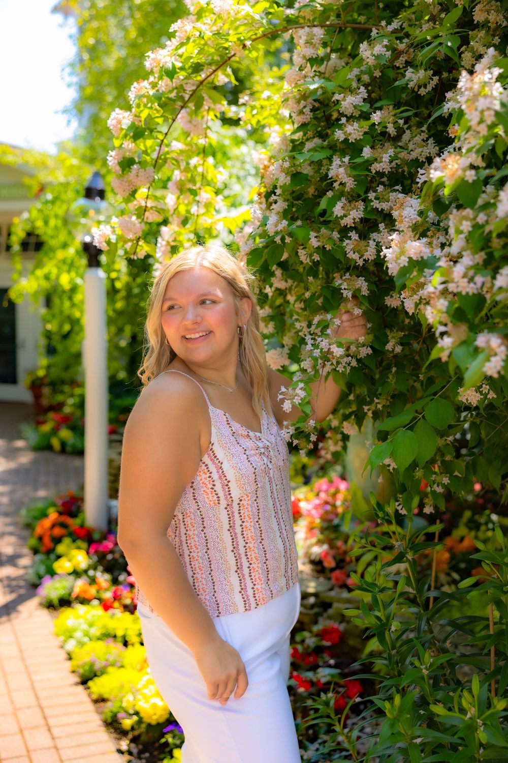 A person wearing a floral crown poses in a lush garden setting with blooming flowers and greenery.