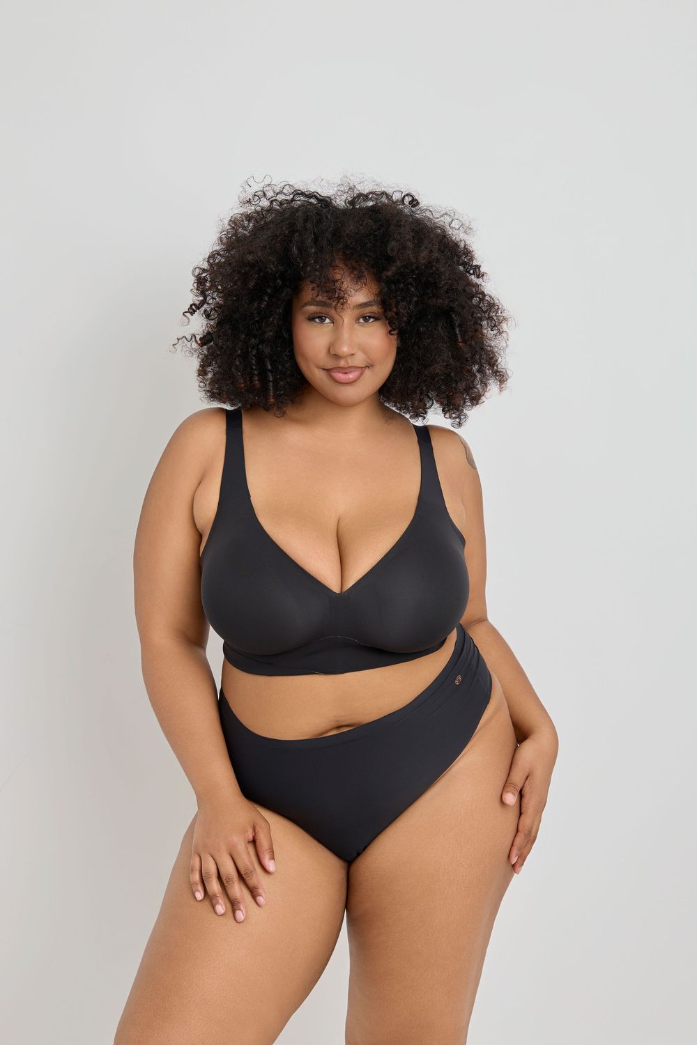 Body positive lingerie model posing in black two-piece set against white background.