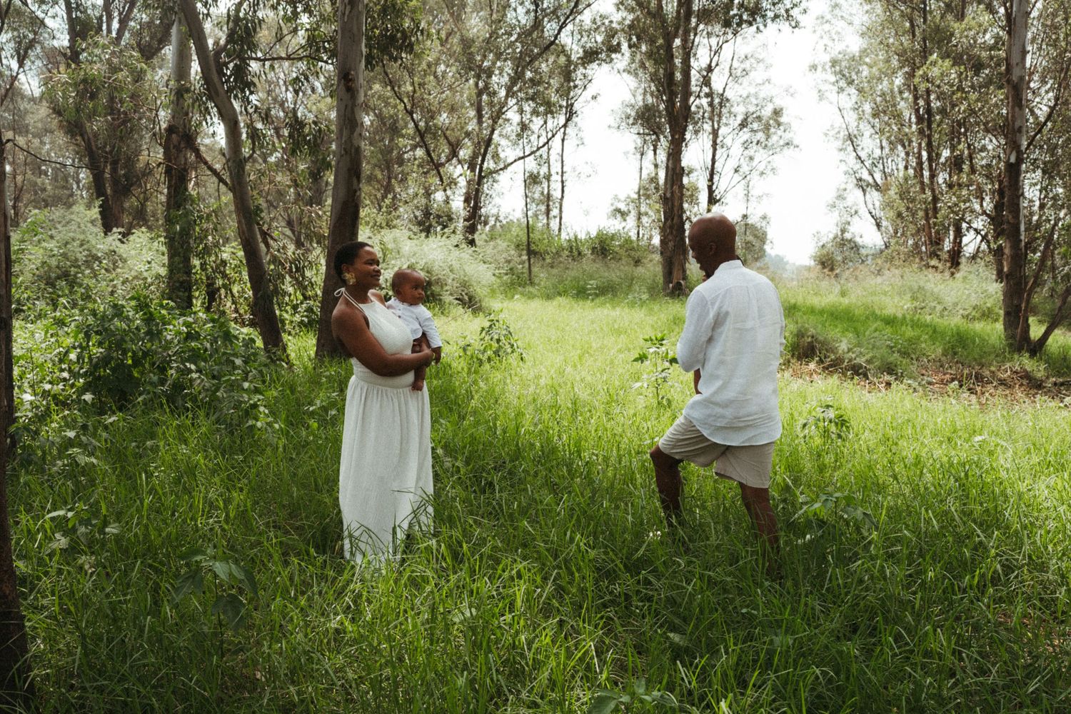 Family wearing white outfits poses together in a lush green outdoor setting with eucalyptus trees.