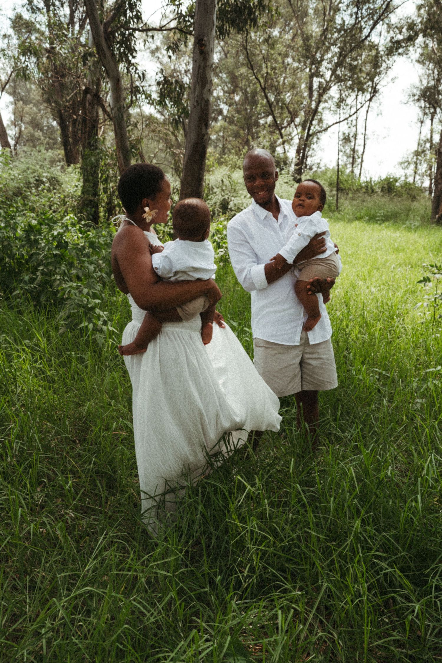 Family in matching white outfits embraces in a natural outdoor setting with tall grass.