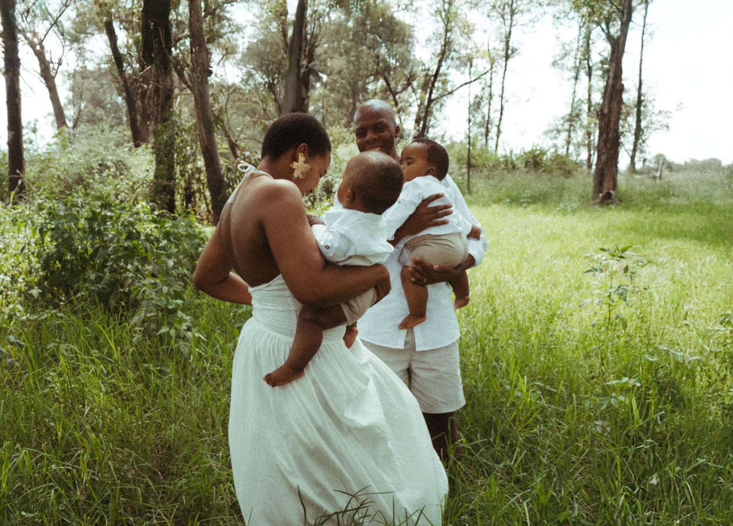 Family group wearing white clothes interacts lovingly in a grassy outdoor setting.