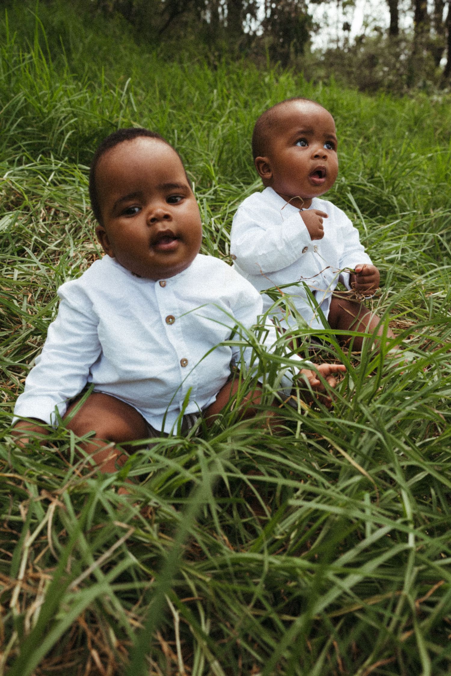 Baby in white shirt sitting in tall grass looking upward with joyful expression.
